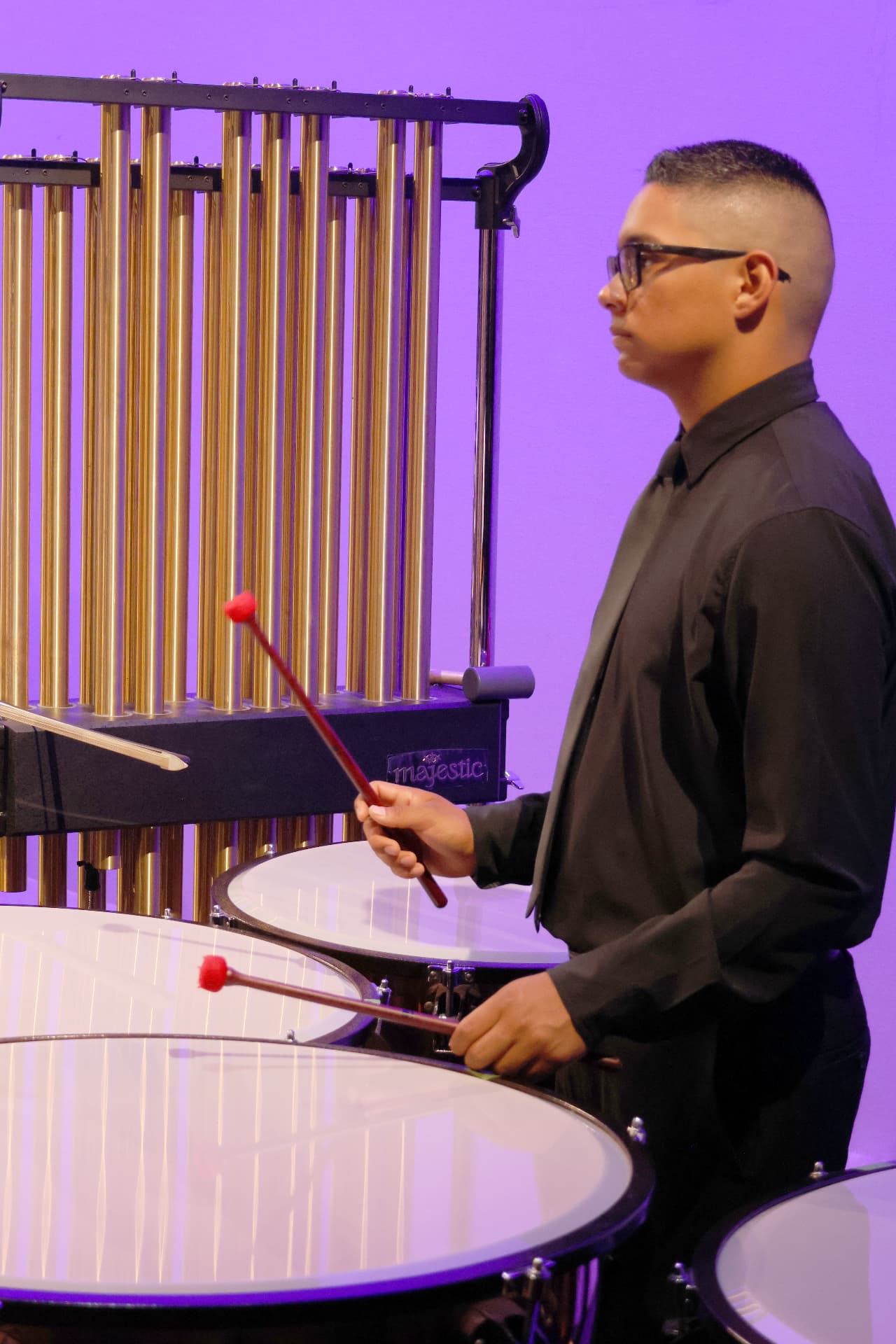 A young percussionist with red mallets at the timpani beside a rack of tubular chimes