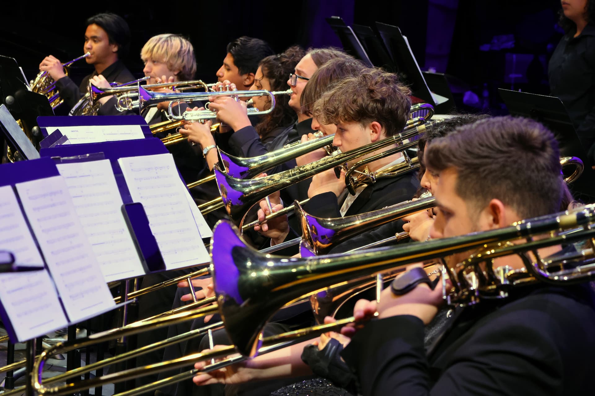A row of trombones with slides extended, trumpets and a French horn seated behind them