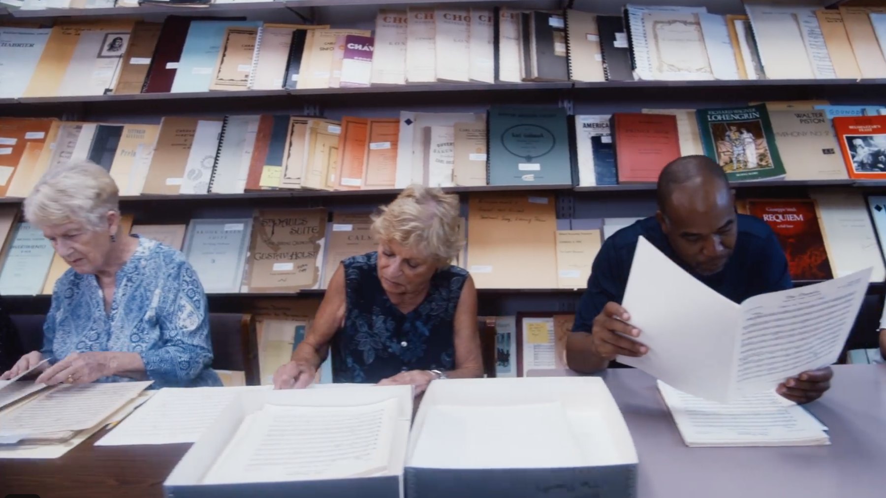 Three people examine full scores at a table before shelves of music