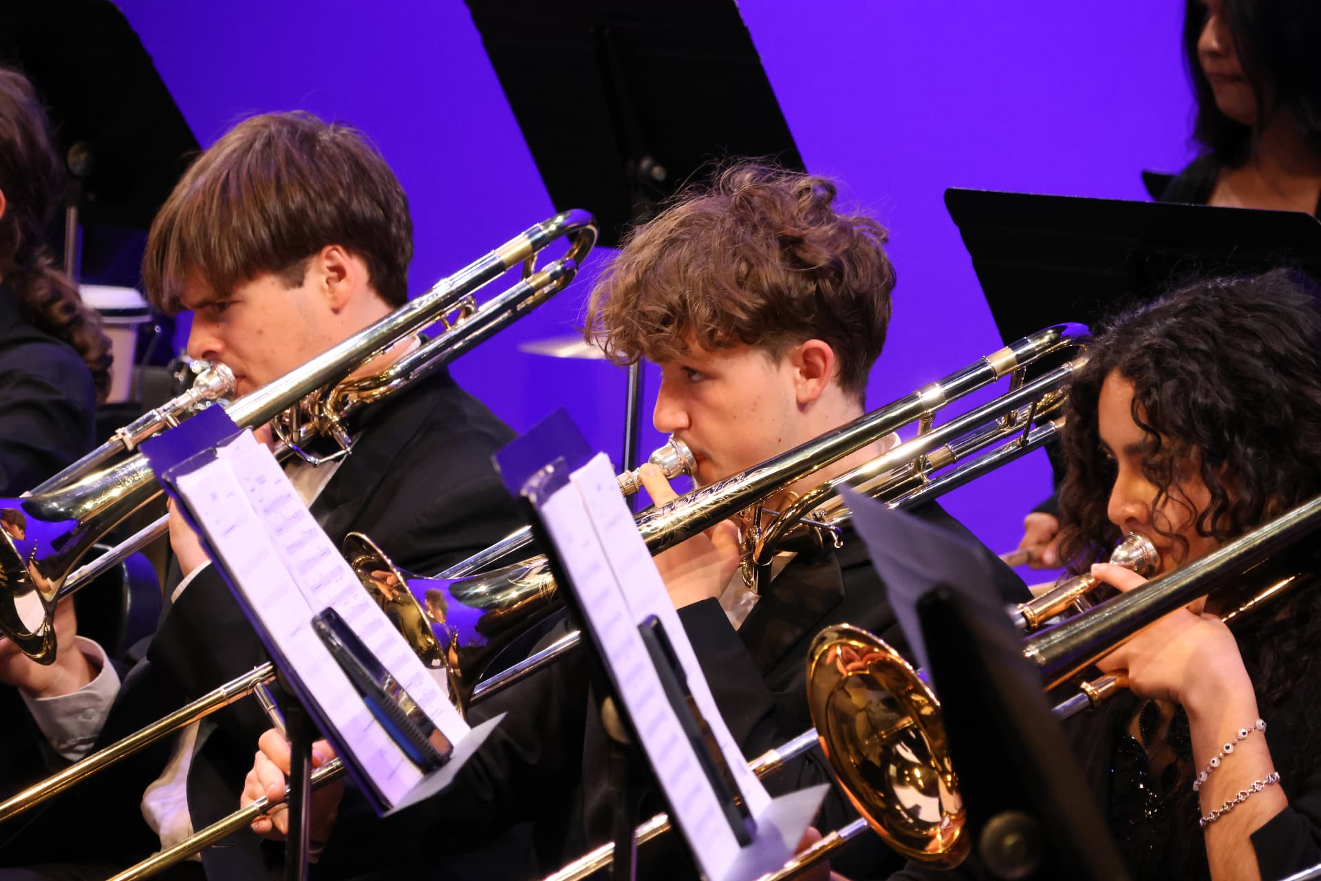 Three young trombone players of the San Bernardino Youth Symphony Orchestra performing in concert black under violet stage light