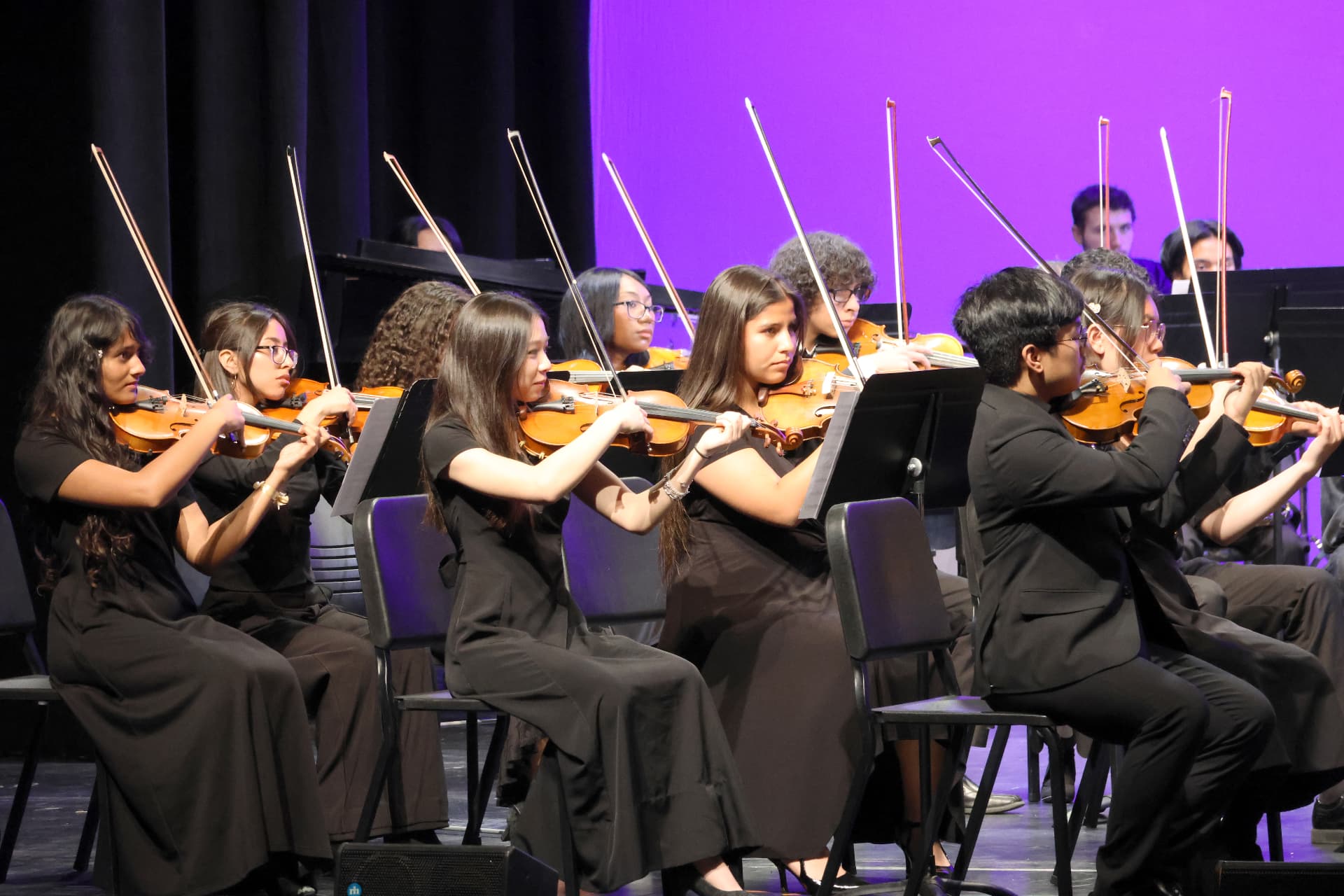 Rows of violinists of the Youth Symphony Orchestra playing with bows raised under violet stage light